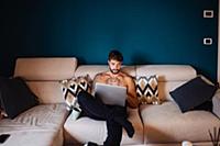 Mid adult man with tattooed chest on sofa using la