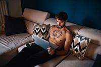 Mid adult man with tattooed chest on sofa looking 