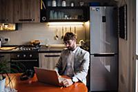 Mid adult man sitting at kitchen table typing on l